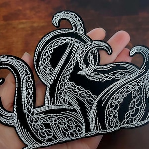 May include: Black octopus patch with white embroidered details. The patch features a detailed design of an octopus with its tentacles curled and flowing. The patch is held in a hand.