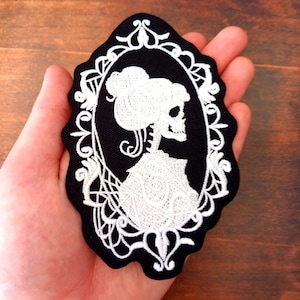 Op de afbeelding: Zwart-wit geborduurde patch met een skelet in een camee-ontwerp. Het skelet draagt een jurk en heeft een gedetailleerd kapsel. De patch heeft een geschulpte rand en wordt in een hand gehouden.