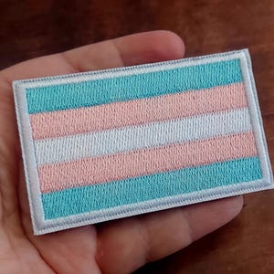 May include: Embroidered rectangular patch featuring the transgender pride flag. The flag design includes horizontal stripes in light blue, pink, and white, with a white border. The patch is likely for clothing or accessories.
