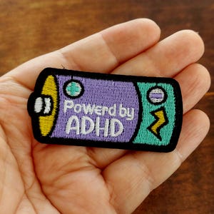 May include: An embroidered patch shaped like a battery with a black border. The left side is yellow, the center is purple with the white text "Powered by ADHD", and the right side is teal with a yellow lightning bolt and a small circular icon.