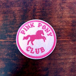 Może przedstawiać: Okrągła, różowo-biała naszywka z haftem z napisem "PINK PONY CLUB" otaczającym różową sylwetkę kucyka. Naszywka znajduje się na ciemnej powierzchni z usłojeniem drewna.
