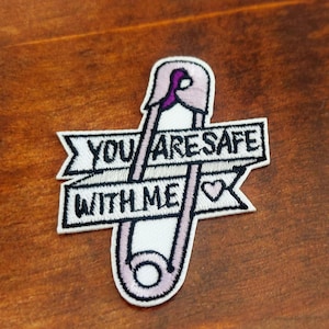May include: Embroidered patch with a pink safety pin design and the text "YOU ARE SAFE WITH ME" in black. The patch includes a small heart symbol. Set against a dark wooden background.