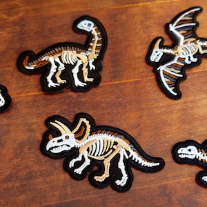 Puede incluir: Un conjunto de parches de esqueletos de dinosaurios. Cada parche presenta un diseño de esqueleto de dinosaurio diferente con detalles en blanco, beige y negro. Los parches están sobre una superficie de madera.