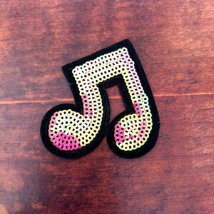 May include: A black sequined music note patch with a yellow and pink gradient design. The patch has a black velvet border. The patch is set against a brown wooden background. Ideal for music-themed crafts.