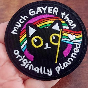 Puede incluir: Parche redondo negro con el texto blanco "much GAYER than originally planned". El parche presenta un gato negro con ojos amarillos y un diseño de bandera arcoíris con un corazón.