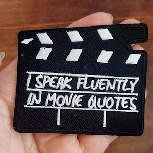 Può includere: Toppa nera a forma di ciak cinematografico con testo ricamato bianco che recita "I SPEAK FLUENTLY IN MOVIE QUOTES". La parte superiore del ciak ha forme rettangolari bianche.