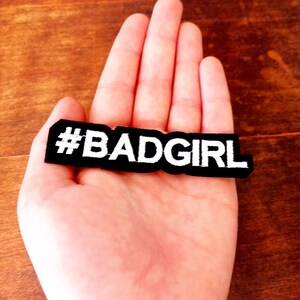 Puede incluir: Un parche rectangular negro con el texto "#BADGIRL" en blanco. El parche se sostiene en una mano abierta sobre una superficie de madera marrón. El parche es un accesorio de ropa.