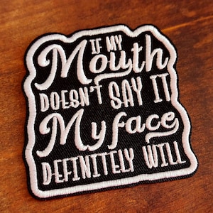 May include: A black embroidered patch with white cursive text that reads "IF MY MOUTH DOESN'T SAY IT MY FACE DEFINITELY WILL". The patch has a white border and is set against a wooden background.