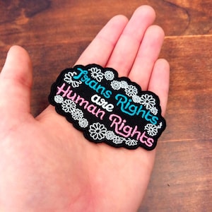 May include: A black embroidered patch with the words "Trans Rights are Human Rights" in white, teal, and pink. The patch is bordered with white daisy-like flowers.