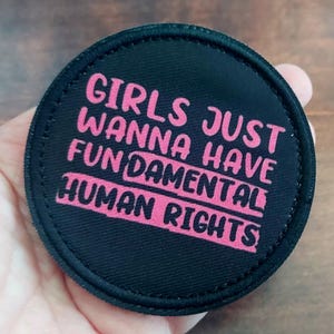 May include: A round, black patch with pink text that reads "GIRLS JUST WANNA HAVE FUNDAMENTAL HUMAN RIGHTS." The patch has a stitched border and is held in a hand.