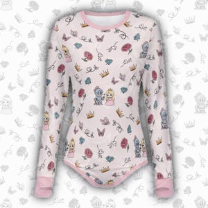 May include: A long-sleeved, white bodysuit with a pink trim. The bodysuit is covered in a pattern of cartoon princesses, knights, crowns, butterflies, roses, and diamonds. The sleeves and neckline are trimmed in pink.