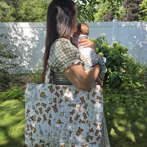 May include: A quilted tote bag featuring a repeating pattern of brown teddy bears, blue lions, and pastel rainbows on a white background. The bag has a square-stitched construction and two shoulder straps, with a small rectangular brand label.