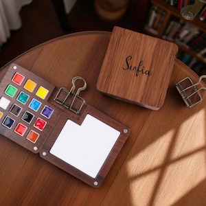 May include: A wooden watercolor palette with a variety of colorful paints, a white mixing area, and a binder clip. The palette is open, revealing the vibrant colors. A wooden box with the name "Sofia" is also present.