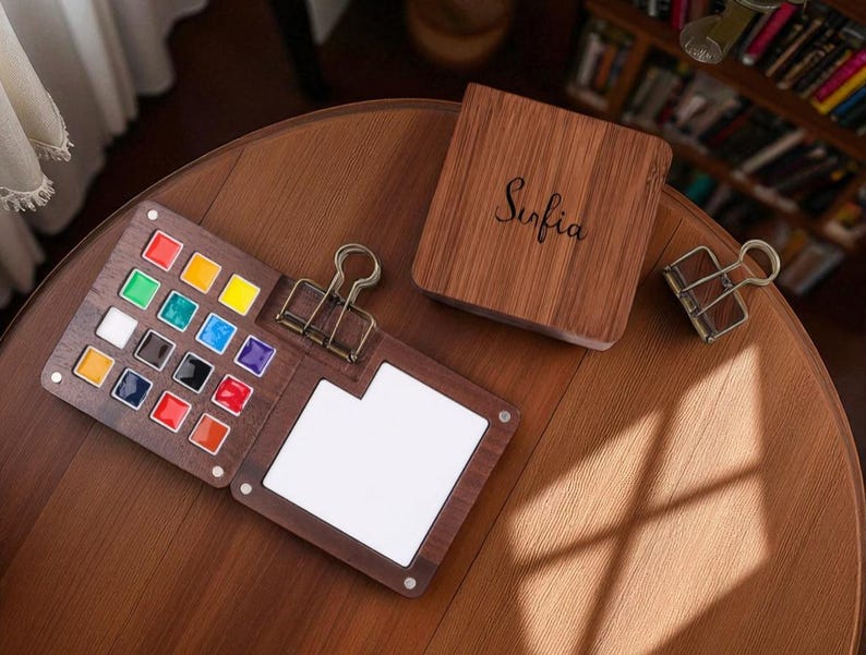 May include: A wooden watercolor palette with a variety of colorful paints, a white mixing area, and a binder clip. The palette is open, revealing the vibrant colors. A wooden box with the name "Sofia" is also present.