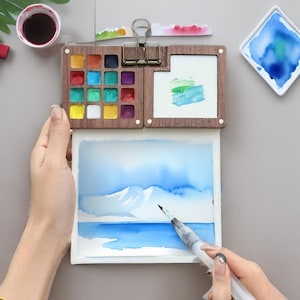 May include: A watercolor painting kit with a wooden palette containing various colors, a small watercolor painting, and a brush. The painting depicts a blue landscape with mountains and water. A small cup of dark liquid is also visible.