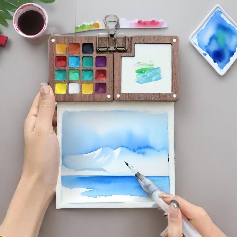 May include: A watercolor painting kit with a wooden palette containing various colors, a small watercolor painting, and a brush. The painting depicts a blue landscape with mountains and water. A small cup of dark liquid is also visible.