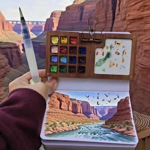 May include: A watercolor painting kit with a wooden palette, a water brush, and a sketchbook. The sketchbook displays a landscape painting of a canyon with a river and birds. The palette contains various colors of watercolor paints.