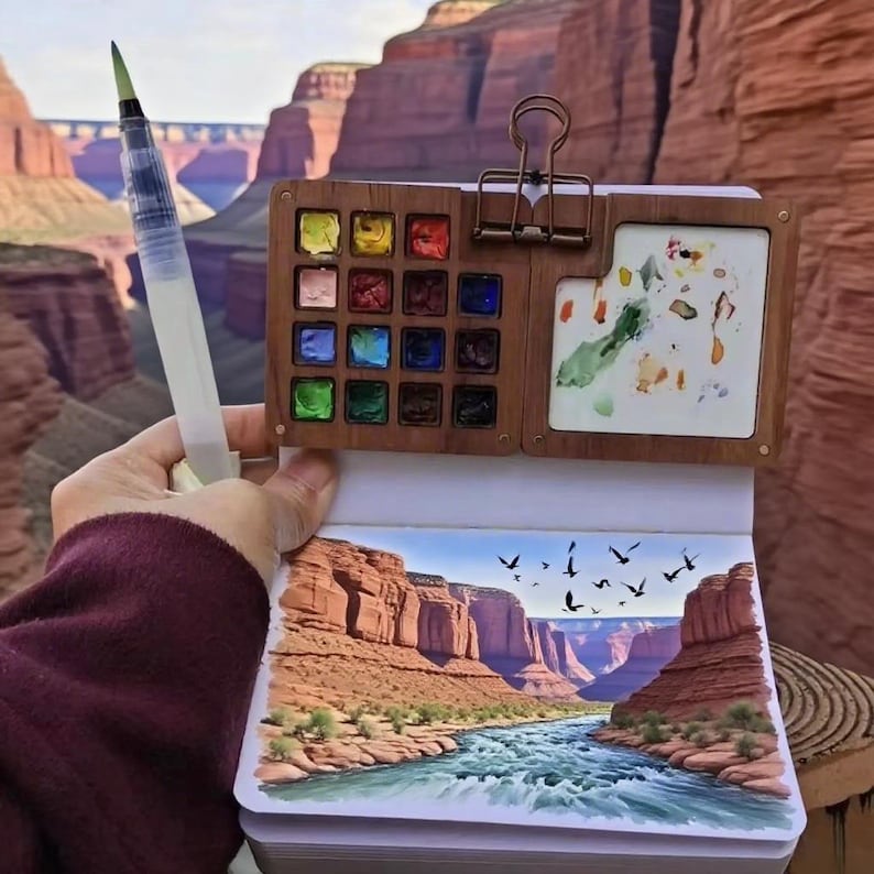 May include: A watercolor painting kit with a wooden palette, a water brush, and a sketchbook. The sketchbook displays a landscape painting of a canyon with a river and birds. The palette contains various colors of watercolor paints.