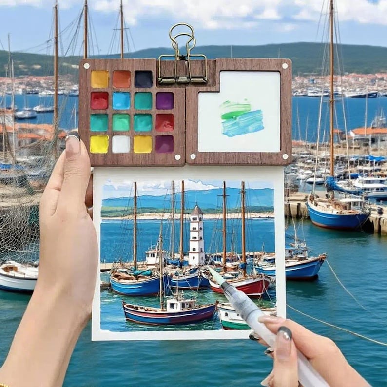 May include: A watercolor paint set with a wooden palette, featuring a variety of vibrant colors. The set includes a watercolor painting of boats in a harbor. A water brush is used to paint the watercolor image.