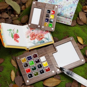Può includere: Palette di acquerelli in legno con vernici colorate e un'area di miscelazione bianca sono esposte. Un taccuino di acquerelli con illustrazioni floreali e di bacche è aperto. È presente anche un pennello ad acqua.