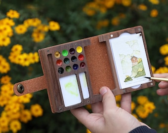 May include: A wooden watercolor palette and sketchbook set. The palette contains a variety of colorful paints, with a small watercolor painting on the right. The set is held open, revealing the contents, with a leather strap closure.