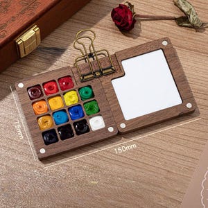 May include: A wooden watercolor paint set with 16 vibrant colors, a mixing area, and a brass clip. The open palette measures 150mm x 75mm. A red rose rests nearby. The set is designed for artists and creatives.