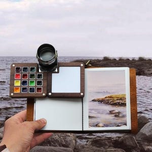 May include: A wooden watercolor palette and sketchbook set. The palette features a selection of colorful paints, a metal water container, and two white mixing areas. The sketchbook displays a watercolor painting of a coastal landscape.