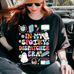 May include: A black t-shirt featuring a colorful graphic with the text "IN MY SPOOKY DISPATCHER ERA" surrounded by illustrations of ghosts, a computer tower, a monitor, a keyboard, headphones, flowers, and small text phrases like "dispatch or treat".
