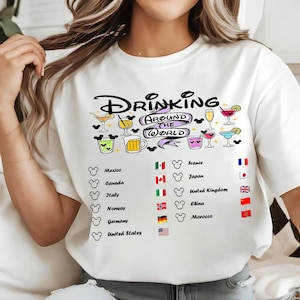 Könnte beinhalten: Ein weißes T-Shirt mit dem Text Drinking Around The World in schwarzer Schreibschrift und einem violetten Banner. Das Design enthält bunte Illustrationen verschiedener Cocktails und Bierkrüge sowie eine Liste von Ländern und deren Flaggen.