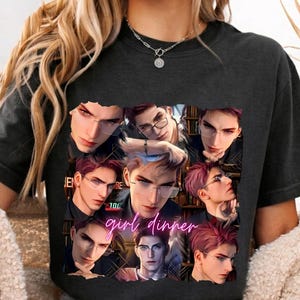 Può includere: Una t-shirt grigio antracite con un collage di diverse illustrazioni in stile anime di una persona con capelli rosso-viola e occhiali. Il testo "girl dinner" è stampato in un carattere corsivo rosa brillante al centro.