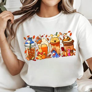 May include: A white short-sleeved t-shirt featuring a graphic print of four beverage containers decorated with autumn-themed illustrations of Eeyore, Winnie the Pooh, Tigger, and Piglet, surrounded by orange and brown fall leaves and pumpkins.