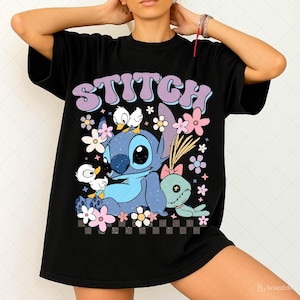 以下が含まれることがあります： 黒いTシャツ。カラフルなスティッチのグラフィックが特徴で、花と小さなアヒルの子に囲まれています。「STITCH」の文字が上部に大きな泡文字で書かれています。デザインには、下部に市松模様が含まれています。