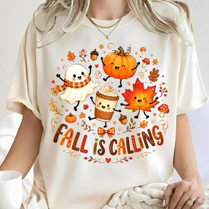 May include: A cream-colored t-shirt featuring a graphic print of a smiling ghost, a pumpkin, a coffee cup, and a maple leaf surrounded by small autumn elements like acorns and mushrooms, with the text "FALL IS CALLING" in brown letters.