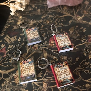 May include: Miniature book keychains featuring the covers of the Harry Potter series. The keychains have silver-colored hoops and are in various colors, including red, green, and gold. The book covers display the title "Harry Potter" and the book's title.