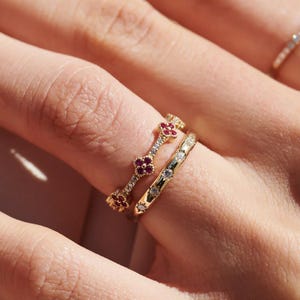 Puede incluir: Dos anillos de oro usados juntos. Un anillo presenta una serie de pequeños motivos florales de cuatro pétalos engastados con piedras rojas y diminutas piedras claras. El segundo anillo es una banda de oro lisa con piedras claras redondas espaciadas.