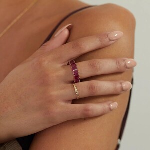 May include: A gold ring featuring a series of rectangular, deep pink gemstones set in a continuous band, worn alongside a thin, textured gold band. The rings are positioned on the fingers, showcasing the vibrant color and the metallic finish.