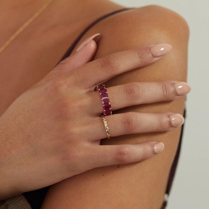 May include: A gold ring featuring a series of rectangular, deep pink gemstones set in a continuous band, worn alongside a thin, textured gold band. The rings are positioned on the fingers, showcasing the vibrant color and the metallic finish.