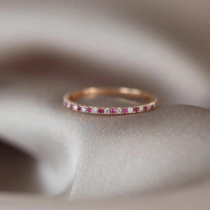 Puede incluir: Un anillo de banda fina de oro con un patrón alterno de pequeños diamantes redondos transparentes de talla brillante y rubíes de color rojo rosado vibrante, engastados a lo largo de toda la circunferencia de la banda metálica en un diseño continuo.