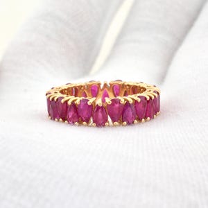 Puede incluir: Un anillo dorado con una fila continua de gemas rojas rubí vibrantes en forma de lágrima. El anillo se presenta sobre un fondo blanco, resaltando el contraste entre el oro y las gemas rojas. El anillo es una pieza de joyería.