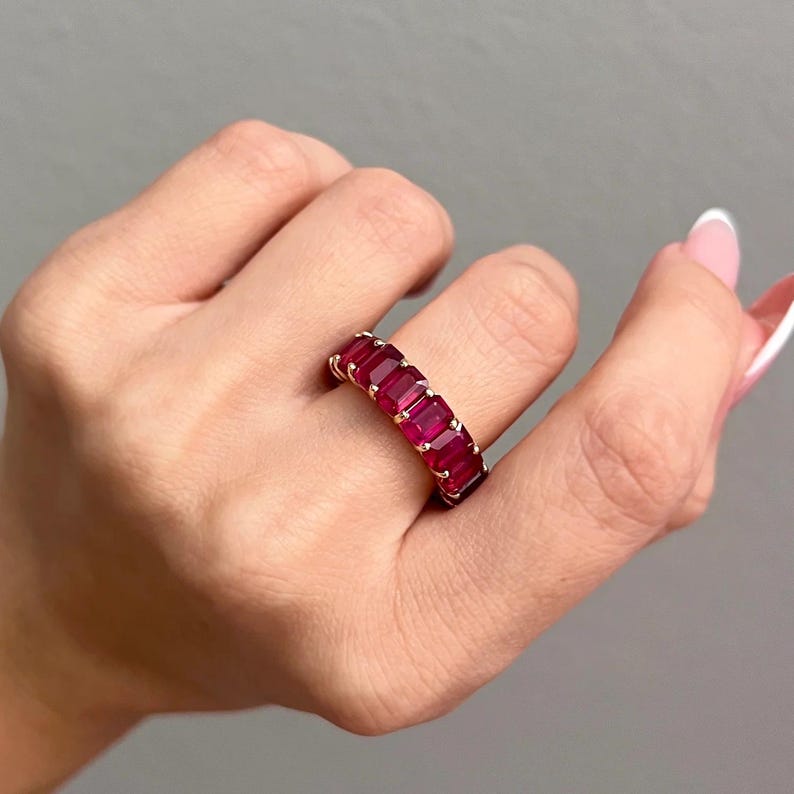May include: A gold band ring featuring a continuous row of vibrant red, emerald-cut gemstones set in a prong setting. The ring encircles the finger, showcasing multiple rectangular-shaped red stones aligned side by side in a polished gold metal frame.