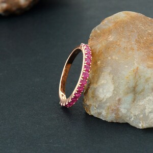 Puede incluir: Un anillo de oro rosa con una banda de peque&ntilde;as gemas rojas vibrantes. El anillo se muestra sobre un fondo oscuro y una roca clara, resaltando el dise&ntilde;o y el color del anillo.