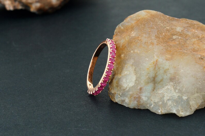 Puede incluir: Un anillo de oro rosa con una banda de peque&ntilde;as gemas rojas vibrantes. El anillo se muestra sobre un fondo oscuro y una roca clara, resaltando el dise&ntilde;o y el color del anillo.