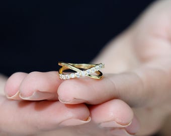 以下が含まれることがあります： ゴールドカラーのクロスデザインのリング。小さな透明な宝石がバンドにセットされています。リングは暗い背景の中で人の手で持たれています。リングのデザインは、交差する2つのバンドが特徴です。