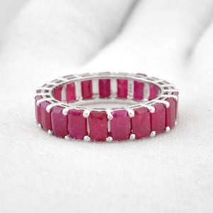 May include: A silver ring with a continuous band of rectangular, deep red gemstones. The ring is set against a white background, highlighting the vibrant color of the stones. The gemstones are evenly spaced around the ring.