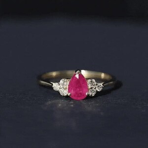 May include: A gold ring featuring a central pear-shaped red ruby gemstone flanked by a cluster of three small round white diamonds on each side. The band is thin and polished, showcasing a classic design with a vibrant, deep pinkish-red stone.