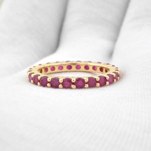 Op de afbeelding: Een gouden ring met een volledige eternity band van ronde, levendige rode edelstenen. De ring is geplaatst tegen een witte achtergrond, waardoor de kleur en het ontwerp van de ring worden benadrukt. De edelstenen zijn gelijkmatig verdeeld.
