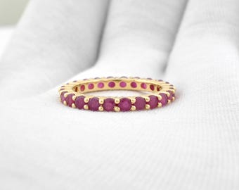 Könnte beinhalten: Ein goldener Ring mit einem durchgehenden Eternity-Band aus runden, leuchtend roten Edelsteinen. Der Ring ist vor einem weißen Hintergrund platziert, der die Farbe und das Design des Rings hervorhebt. Die Edelsteine sind gleichmäßig angeordnet.