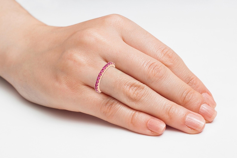 Puede incluir: Un anillo de oro rosa con una banda de peque&ntilde;as y vibrantes piedras preciosas rosas. El anillo se lleva en el dedo de una persona, mostrando el delicado dise&ntilde;o y color. El anillo es una joya.