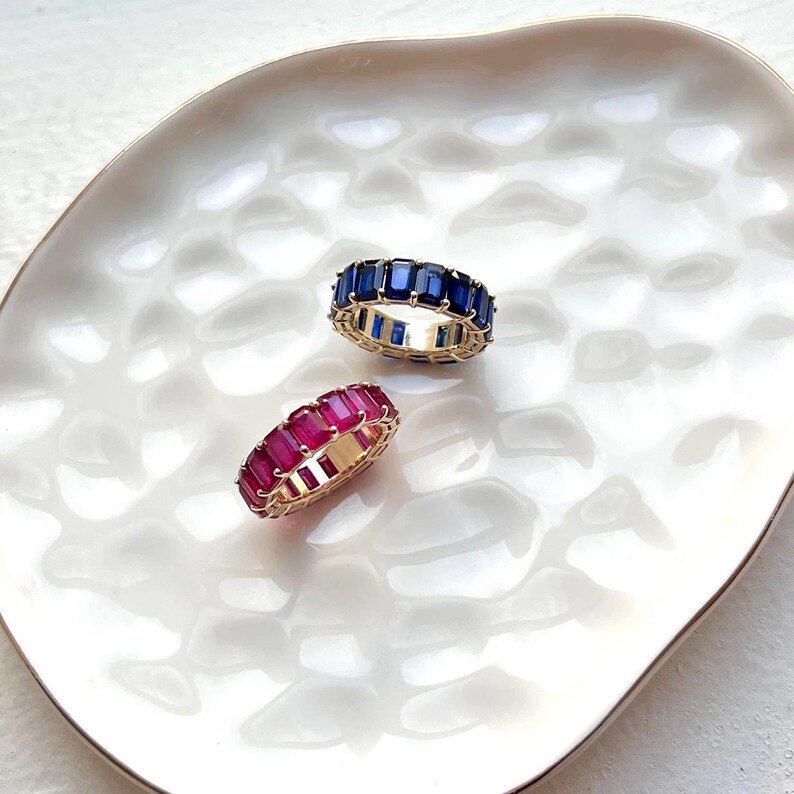 May include: Two eternity bands featuring baguette-cut stones set in gold-toned metal. One ring displays deep blue stones, while the other ring features vibrant magenta-pink stones. Both bands are circular with a continuous row of gemstones.