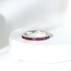 May include: A silver-toned band ring featuring a continuous row of rectangular, baguette-cut deep red stones set horizontally. The metal edges of the ring are detailed with a fine, beaded milgrain texture that frames the vibrant red gemstones.